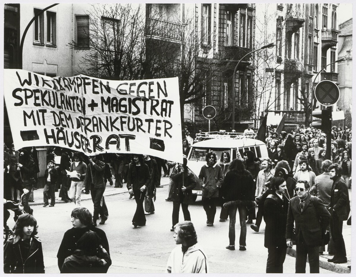 Ein Demo zieht vor einem Altbau vorbei, aus dem Transparente hängen.